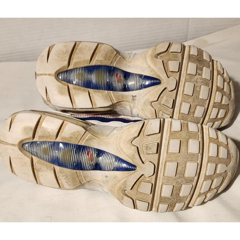 Puede incluir: Par de zapatillas deportivas blancas y azules, vistas desde las suelas. Las suelas muestran un patr&oacute;n texturizado y desgastado con detalles azules y rojos. Las zapatillas parecen usadas.