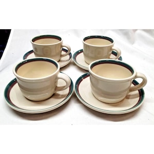 Può includere: Quattro tazze da tè e piattini in ceramica bianco sporco. Ogni tazza ha un manico ed è posizionata su un piattino abbinato. I bordi delle tazze e dei piattini sono rifiniti con una fascia verde e bordeaux. Le tazze e i piattini sono disposti su una superficie bianca.