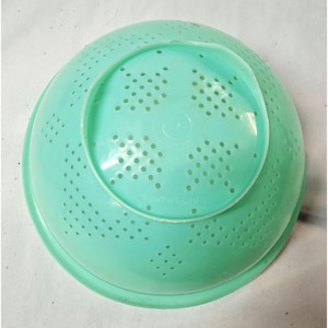 Op de afbeelding: Een ronde, mintgroene plastic vergiet met een verhoogde cirkelvormige voet en een patroon van kleine afvoergaten gerangschikt in ruitvormige clusters over het oppervlak. Het woord Tupperware is in het midden van de voet gegraveerd.