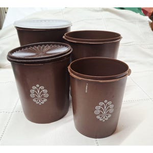 Peut inclure: Un ensemble de quatre boîtes de rangement cylindriques marron avec couvercles assortis. Deux boîtes présentent un motif floral circulaire blanc sur le devant. Les couvercles sont marron avec un motif en rayons ou blanc translucide.