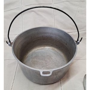 May include: A round, silver-colored metal cooking pot with a smooth interior and a matte exterior finish. The pot features a thin, black, arched wire handle attached to two side lugs and a small, integrated handle loop on the front rim.