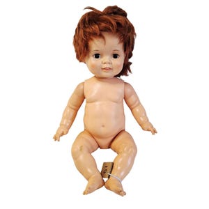 May include: A vintage baby doll with red hair and blue eyes, seated with arms and legs extended. The doll has a tag attached to its leg. The doll is made of a light-colored material.
