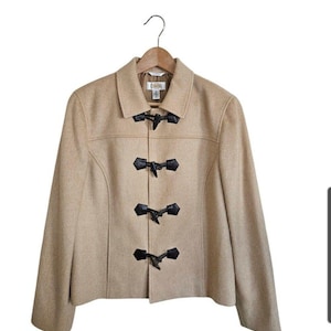May include: A tan wool-blend jacket featuring a classic collar and four black toggle closures with leather-like tabs down the front. The jacket has long sleeves, a straight hemline, and is displayed on a wooden hanger against a white background.