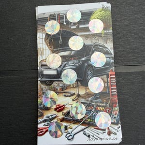 May include: A rectangular sticker sheet featuring a detailed illustration of a black car with its hood open inside a workshop, surrounded by various tools and equipment. The sheet is decorated with ten circular holographic stickers.