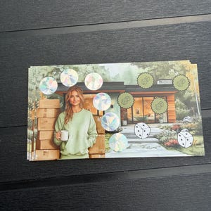 May include: A rectangular sticker sheet featuring a printed illustration of a person in a light green sweatshirt holding a white mug next to stacked cardboard boxes in front of a modern house, accented with holographic circles and dice icons.