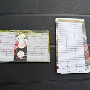 May include: Two stacks of rectangular paper cards featuring a printed grid with columns labeled Datum, +/-, and Summe. The cards display a background image of a red sports car and include three circular holographic stickers on the horizontal stack.