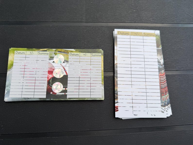 May include: Two stacks of rectangular paper cards featuring a printed grid with columns labeled Datum, +/-, and Summe. The cards display a background image of a red sports car and include three circular holographic stickers on the horizontal stack.