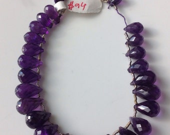 Puede incluir: Un collar de cuentas de amatista facetadas en forma de lágrima, de color púrpura oscuro. Las piedras están ensartadas en un alambre, con una borla y una etiqueta con el número "#94".