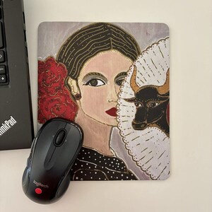 Pode incluir: Tapete de rato retangular com um retrato pintado de uma mulher com rosas vermelhas e um touro preto atrás de um leque branco. A mulher tem batom vermelho e usa um vestido preto com bolinhas. Um rato Logitech preto está em primeiro plano.
