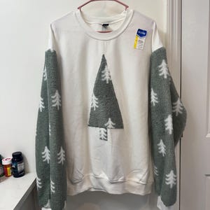 May include: White sweatshirt with a large gray tree design on the front. The sleeves are a soft gray material with white tree patterns. The sweatshirt is hanging on a pink hanger.