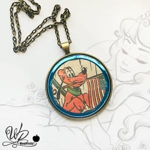 Puede incluir: Un collar con colgante redondo con un perro de dibujos animados sosteniendo una bandera estadounidense. El colgante tiene un borde azul y una cadena de color bronce. El perro es rojo y lleva una bufanda verde.