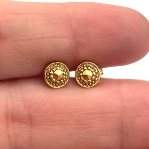 May include: A pair of small, gold-colored stud earrings. Each earring has a circular design with a textured border and a central, raised geometric pattern. The earrings are displayed on a light background.