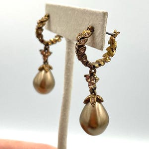 May include: A pair of gold-toned hoop earrings with a floral design and teardrop-shaped pearl pendants. The earrings are displayed on a neutral-colored earring stand. The earrings have a vintage or antique aesthetic.