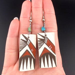 May include: Handmade rectangular earrings with a white base, featuring a geometric design in red, black, and white. Each earring has a silver hook and a small turquoise or green bead. The earrings are held in a hand.