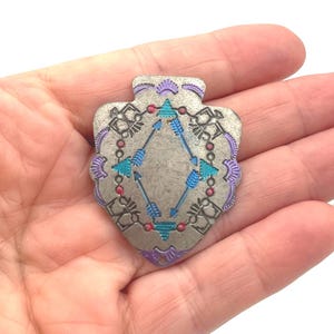 May include: A silver-toned metal pendant with a shield shape. The pendant features a central diamond design with blue arrows and teal and red accents. The edges are adorned with purple and teal details.