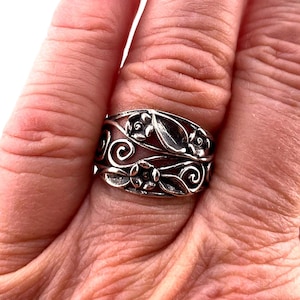 May include: A wide silver band ring featuring an intricate openwork design of embossed floral motifs and swirling vine patterns. The metal has a darkened, oxidized finish in the recessed areas to highlight the detailed relief of the flowers.