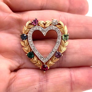 May include: A heart-shaped brooch featuring a gold-tone metal frame with a leaf motif. The inner heart border is lined with small clear crystals, while the outer edge is accented with six round, multi-colored gemstones in pink, green, and blue.