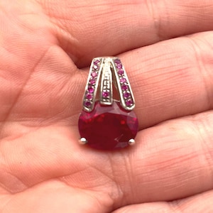 Può includere: Un pendente in argento con una grande gemma di rubino taglio ovale. Il rubino è di colore rosso intenso ed è accentuato da gemme rosa più piccole incastonate nella cauzione d'argento. Il pendente è progettato per essere indossato come collana.