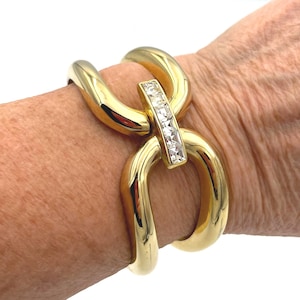 Op de afbeelding: Een goudkleurige armband met een schakelkettingontwerp. De armband heeft een rechthoekige staaf met heldere kristallen. De armband wordt om de pols gedragen.