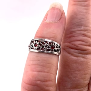 May include: A silver-toned band ring featuring an openwork filigree design of scrolling vines and leaves. The metal surface has a darkened, oxidized finish in the recessed areas, and two small, round, deep red stones are set within the floral motifs.