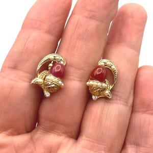 Puede incluir: Un par de pendientes dorados con forma de bellota, con detalles de piedras preciosas rojas y pequeñas perlas blancas. Los pendientes tienen un diseño detallado con una pequeña piedra similar a un diamante en la base de cada bellota.