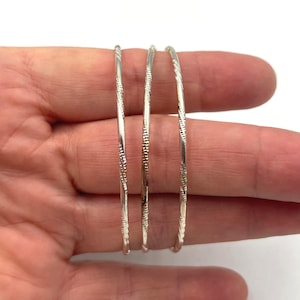 May include: Three silver bangles with textured designs are displayed. The bangles are thin and appear to be made of metal. The background is plain white, highlighting the jewelry.
