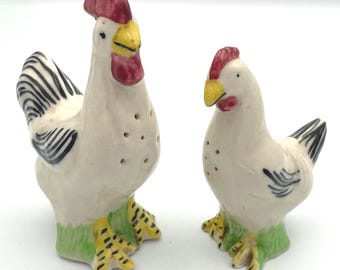 May include: A pair of ceramic salt and pepper shakers in the shape of a rooster and hen. The rooster has a red comb and wattle, yellow beak, and black and white tail feathers. The hen has a red comb and yellow beak. Both are on green bases.