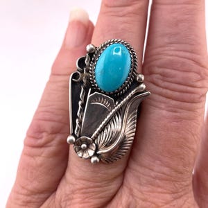 May include: A sterling silver ring featuring a central oval turquoise stone surrounded by a twisted wire border. The setting includes decorative metalwork with a leaf motif, a small flower accent, and etched line details on the silver surface.