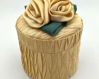 May include: A small, round, gold-colored box with a pleated texture. The lid is adorned with two fabric roses in a matching gold hue, set atop green fabric leaves. A decorative storage container.