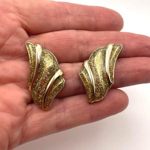 Puede incluir: Un par de pendientes dorados con clip. Cada pendiente tiene una forma curva, similar a un abanico, con una superficie texturizada y brillante en tonos dorados y bronce. Los pendientes se sostienen en una mano sobre un fondo blanco.