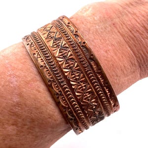 Pode incluir: Uma pulseira de cobre larga tipo bracelete com padrões estampados intrincados, incluindo motivos de estrelas, texturas lineares e bordas recortadas. O metal tem um acabamento polido quente com áreas rebaixadas escuras que destacam os detalhes.