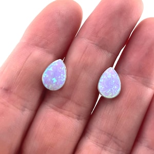 May include: A pair of teardrop-shaped stud earrings featuring iridescent light purple opal stones set in a thin silver-toned metal frame. The stones display a speckled pattern with flashes of blue and pink light across their smooth surfaces.