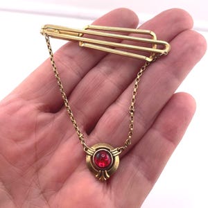 May include: A gold-tone tie bar featuring a triple-bar design connected by a delicate chain to a circular pendant. The pendant is centered with a round, translucent red stone set in a gold-tone frame with decorative metal detailing at the base.