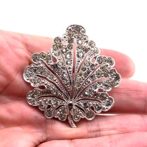 May include: A silver-toned metal brooch shaped like a leaf, featuring an intricate openwork design filled with numerous small, clear, sparkling rhinestones set throughout the entire surface of the leaf-shaped frame.