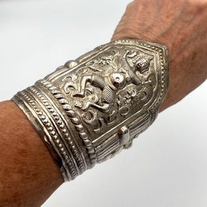 May include: A silver-colored cuff bracelet with intricate relief carvings. The bracelet features a central panel with a detailed figure and floral patterns, surrounded by decorative borders. The cuff is wide and wraps around the wrist.