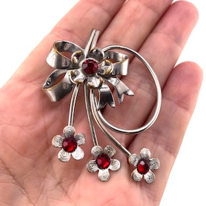 May include: A silver-toned metal brooch featuring a ribbon bow design and three dangling flower charms. Each flower is centered with a small, round, faceted red gemstone. The brooch has a polished finish with curved metal stems and a circular loop.