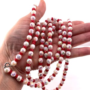 May include: A long necklace featuring alternating iridescent white faceted beads and small solid red cylindrical beads. The strand is strung with a repeating pattern of these two bead types and includes a silver-toned metal clasp at one end.