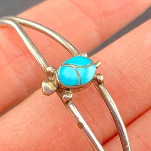 May include: A silver-toned bangle bracelet featuring a turquoise turtle design. The turtle has a bright blue stone center and is flanked by small silver accents. The bracelet's slender, curved design adds a delicate touch to the jewelry.
