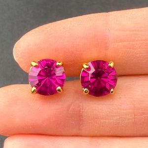 May include: A pair of stud earrings featuring large, round, magenta-colored gemstones set in gold-colored metal. The earrings are displayed against a neutral background.