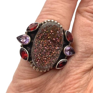 May include: A silver ring featuring a large, oval-shaped, sparkling druzy stone in shades of brown and red. The ring is accented with smaller, teardrop-shaped red and purple gemstones around the central stone. The ring is on a hand.