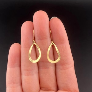 May include: A pair of gold-colored teardrop earrings. The earrings are held between fingers against a dark background. Each earring has a simple, open teardrop shape suspended from a small hook.