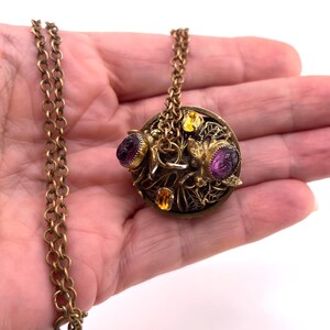 May include: A round brass pendant necklace featuring a detailed filigree base adorned with two purple carved scarab-style gemstones and two smaller yellow oval-cut stones. The pendant is attached to a matching brass link chain with a circular clasp.