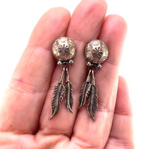 May include: A pair of silver-toned earrings featuring a round, stamped dome top with a sunburst pattern. Each earring has two dangling, detailed feather charms attached to the base of the dome by small metal loops, creating a layered, textured look.