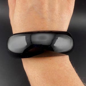 May include: A glossy, black bracelet with a rectangular shape. The bracelet is worn on a wrist, reflecting light. The jewelry piece is a bold accessory.