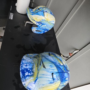 May include: Two blue and yellow marbled helmets are displayed on a black surface. The helmets have a unique design with a mix of blue, yellow, and white colors. The helmets appear to be for sports or recreational use.
