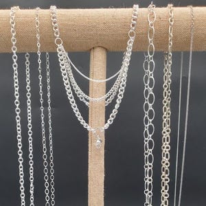 May include: A display of various silver necklaces hanging from a tan jewelry stand. The necklaces feature different chain styles, including delicate strands and chunky links. The background is a solid black, highlighting the metallic shine of the jewelry.