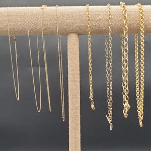 May include: A selection of gold-coloured necklaces displayed on a tan fabric-covered jewellery stand. The necklaces showcase diverse chain styles, from fine strands to beaded and chunky link designs. The backdrop is a plain dark grey.
