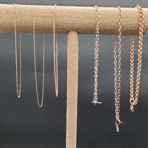 May include: A display of rose gold necklaces on a tan jewelry stand. The necklaces vary in style, including delicate chains, beaded strands, and chunkier link designs. The background is a solid dark gray.