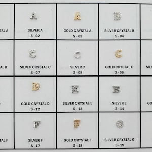 May include: A grid of small, decorative letter charms in silver and gold tones. The letters A, B, C, D, E, F, and G are displayed in various styles, some with crystal accents. Each charm is labeled with its material and a unique identifier.