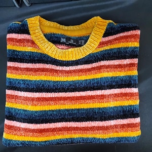 May include: A folded, multi-colored striped sweater. The sweater features horizontal stripes in shades of red, yellow, blue, and black. The collar is a solid yellow. The sweater is made of a soft, textured material.